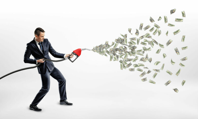 A smiling businessman holding a gas nozzle while many dollar bills are flying out of it instead of petrol.