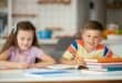 Two children sitting at a table doing homework, with notebooks, pencils and colourful books in front of them.