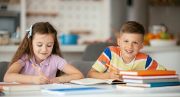 Two children sitting at a table doing homework, with notebooks, pencils and colourful books in front of them.
