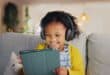 Young girl wearing headphones, smiling while using a tablet on a couch with a soft toy beside her.