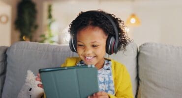 Young girl wearing headphones, smiling while using a tablet on a couch with a soft toy beside her.