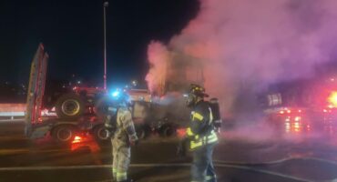 Two firefighters in full protective gear stand near the smouldering vehicle transporter truck on the N1 near Beyers Naudé Drive, as thick smoke rises into the night sky. Photo: Supplied