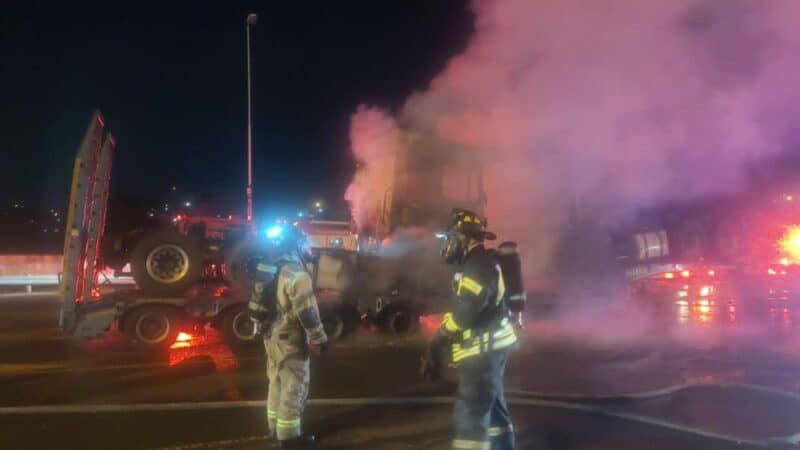 Two firefighters in full protective gear stand near the smouldering vehicle transporter truck on the N1 near Beyers Naudé Drive, as thick smoke rises into the night sky. Photo: Supplied