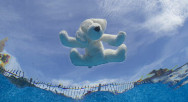 A white stuffed animal, resembling a dog, floats on a clear blue pool surface. The sky above is bright with wispy clouds, creating a serene scene.