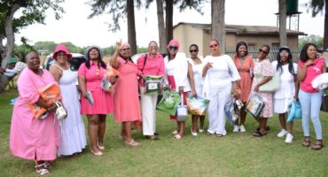 A group of women from Sekhukhune District Municipality in bright pink and white outfits stand on grass, smiling and holding gift bags. Tall trees and a small building are in the background.