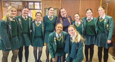A group of nine young women, smiling widely, pose in a school setting. Eight are in green uniforms with the Hoërskool Ben Viljoen badges, suggesting unity and achievement.