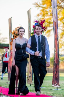 A couple walks arm-in-arm on a red carpet during a matric farewell event. The woman wears an elegant black gown, and the man is in a vest and tie. Floral arrangements are in the background.