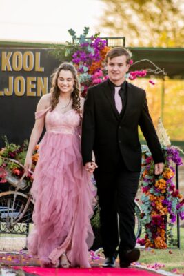 A young couple walks hand-in-hand down a red carpet at a matric farewell. The woman wears a flowing pink gown, while the man is in a black suit with a pink tie. They are surrounded by vibrant floral decorations. The mood is joyful and festive, with a sunset backdrop adding warmth to the scene.