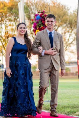 A smiling couple walks arm-in-arm on a red carpet during a matric farewell event. The woman wears a blue gown with floral details, the man in a beige suit. Bright flowers in the background.