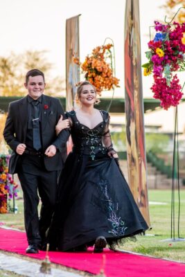 A young couple, dressed in elegant black attire, walks arm-in-arm on a red carpet at a matric farewell event. They are surrounded by colorful floral arrangements, smiling joyfully.