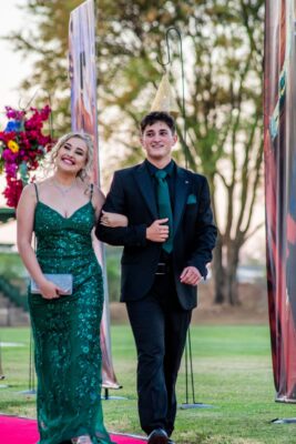 Young couple dressed formally, walking on red carpet outdoors during a matric farewell event. Woman in sparkling green gown, man in dark suit with green tie, both smiling warmly.
