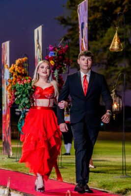 A woman in a vibrant red dress holds red roses, walking down a red carpet alongside a man in a black suit with a red tie during a matric farewell event. Colorful floral arrangements and artwork line the path, set against a twilight sky, conveying a festive and elegant atmosphere.