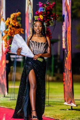 A woman in a sparkling black gown holds a bouquet while walking a red carpet lined with vibrant floral arrangements at a matric farewell event. The scene is evening and elegant.
