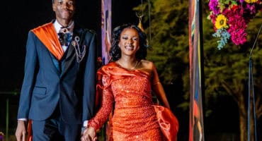 A young couple stands confidently, dressed elegantly for an elegant matric farewell event. The woman wears a sparkling orange gown, and the man is in a black suit with an orange sash. They are smiling, surrounded by floral decorations, with trees visible in the background.