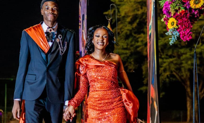 A young couple stands confidently, dressed elegantly for an elegant matric farewell event. The woman wears a sparkling orange gown, and the man is in a black suit with an orange sash. They are smiling, surrounded by floral decorations, with trees visible in the background.