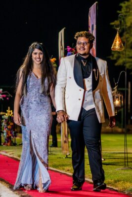 A smiling couple walks hand in hand on a red carpet at night during a matric farewell event. She wears a sparkly silver dress; he wears a white tuxedo jacket. Festive and elegant mood.