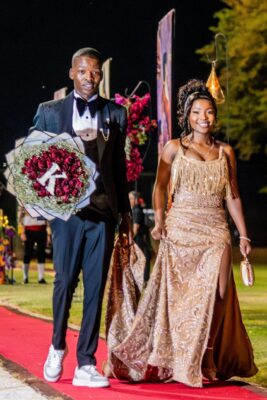 A smiling couple walks on a red carpet at night during a matric farewell event. The man holds a bouquet of red roses and wears a black tuxedo with white sneakers. The woman wears a gold, fringed gown and carries a small clutch. Festive, elegant atmosphere.