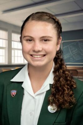 Smiling student wearing a green blazer with a school badge, standing in a classroom with a blackboard and windows. The atmosphere is cheerful and studious.