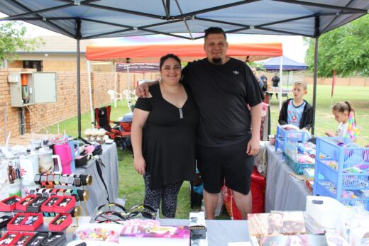 A smiling couple stands under a canopy at an outdoor market stall, displaying various products. The scene is lively with colorful tents in the background.