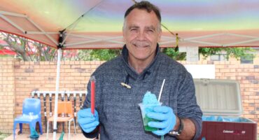 Man smiling under a tent, holding a blue shaved ice and a red popsicle. He's wearing gloves and a sweater, with coolers and bottles nearby.
