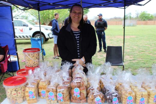 A smiling woman stands behind a table with various packaged cookies under a blue canopy at an outdoor market. A white vehicle and people are visible in the background.