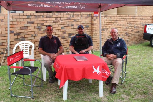 Three men sit under a Loskop Alarms branded canopy at a red table with a Loskop Alarms logo. The setting is outdoors on grass with a brick wall background, creating a friendly, professional atmosphere.