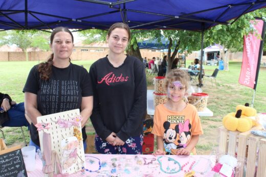 Three people stand behind a craft stall at an outdoor market. They are under a blue canopy with handmade jewelry and plush items displayed. The mood is cheerful.