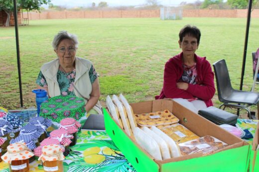 Two women sitting at an outdoor market stall with homemade preserves and baked goods displayed on a colorful tablecloth, conveying a welcoming and friendly atmosphere.