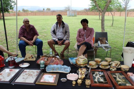 Three people sit under a tent at a flea market, smiling and relaxed. A table in front displays various vintage items: ceramics, framed art, and decorative pieces. The background shows a lawn with trees and a brick wall.