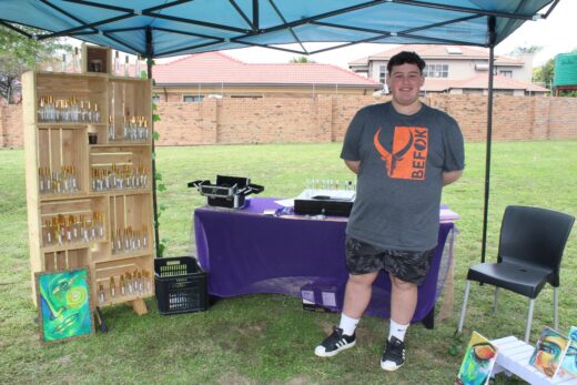 Man smiling under a blue canopy at an outdoor market stall. Shelves with bottles of perfume and vibrant paintings are displayed on a purple-covered table.