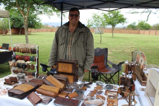 Smiling vendor stands beside a market stall displaying handcrafted leather goods on a table. Green park setting with trees, under a canopy. Casual and inviting ambiance.