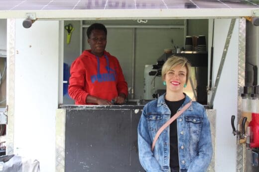 A woman in a denim jacket smiles in front of a food truck while another person in a red hoodie stands inside. This food truck is selling coffee and various other hot and cold drinks. The mood is friendly and inviting.
