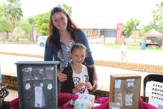 A smiling woman and child stand behind a craft stall displaying jewelry and crafts outdoors. Palm trees and buildings are in the background, conveying a cheerful market atmosphere.