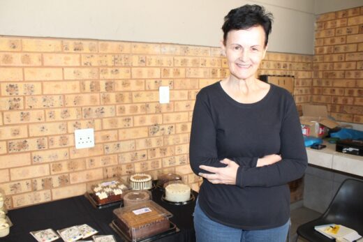 A smiling woman stands in front of a table with various cakes and baked goods, against a brick wall. The scene conveys a warm and inviting atmosphere.