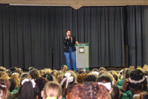 A speaker, Mel die Storieverteller, stands on a stage in front of a dark curtain, addressing an attentive audience of students. The atmosphere is focused and engaged.