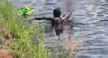 A person swimming near a grassy edge of an irrigation canal, holding a blurred object in his right hand and reaching for the edge of the canal with his left. The scene is calm, with gentle ripples on the water.