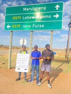 Three men stand under a green road sign with directions to Marulaneng, Lebowakgomo, and Jane Furse. One holds a sign, another has a walking stick. It's sunny.