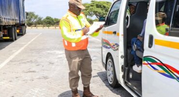 Traffic officer in a high-visibility vest checks paperwork beside a parked van with colorful stripes. The scene is calm, with blue skies and trees.