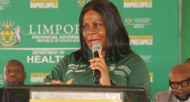 A woman in a green shirt speaks into a microphone at a podium against a backdrop reading "Limpopo Department of Health." The setting is formal and focused.