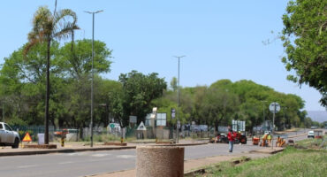 A road with a few vehicles and construction barriers, bordered by green trees under a clear blue sky. A person in red walks on the sidewalk. Calm atmosphere.
