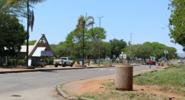 Road maintenance work is evident. A quiet street scene featuring parked vehicles near a triangular entrance sign. Trees and greenery line the road on a sunny day, creating a calm atmosphere.