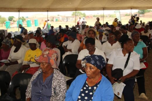 A large group of people sits under a tent in a rural setting, mostly women wearing colorful hats and clothing, attentively listening. The atmosphere is focused.