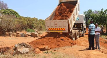 A dump truck unloads a large pile of red soil where a huge hole was on a dirt pathway in Sephaku Village, with two men conversing nearby amid trees and bushes under a clear blue sky.
