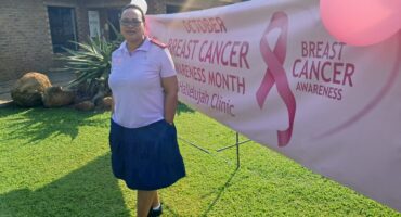 A woman in a pink shirt stands beside a "Breast Cancer Awareness Month" banner with a pink ribbon, on a sunny day with grass and a building behind.