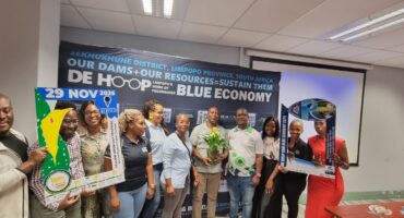 A diverse group of people stands smiling in a room with a banner about water conservation. They hold photo frames and a plant, creating a positive, hopeful tone.