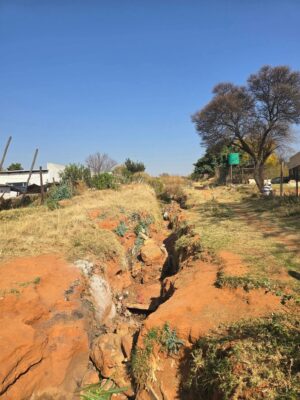 Eroded path with a deep red soil trench under a clear blue sky in Sephaku Village. Sparse grass and scattered trees. The image conveys a sense of neglect. 