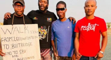 Four men stand outdoors in casual clothing, holding a sign. The sign mentions a walk from Leewfontein to Beitbridge, protesting lawlessness. The mood is serious and determined.