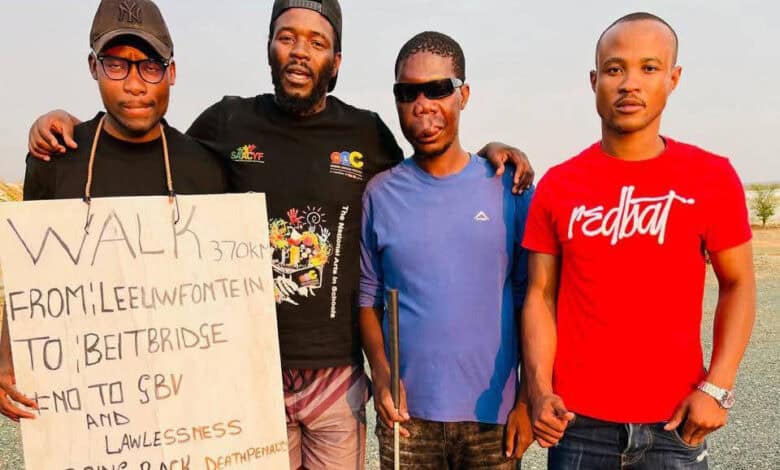 Four men stand outdoors in casual clothing, holding a sign. The sign mentions a walk from Leewfontein to Beitbridge, protesting lawlessness. The mood is serious and determined.