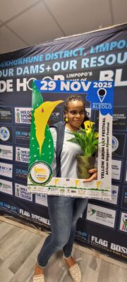 A woman smiles while holding a potted plant, framed by a promotional cutout for the Yellow Arum Lily Festival on November 29, 2025, in front of a sponsor backdrop.
