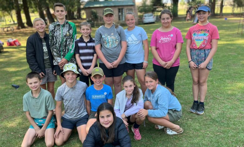 A group of smiling teenagers stands and kneels on grass in a park. They're casually dressed in colorful t-shirts and shorts, exuding a joyful, relaxed vibe.